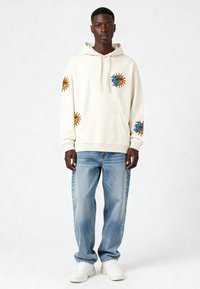 Felpa crema con motivi di sole e luna in arancione e blu, in materiale cotone, tasca frontale, abbinata a jeans azzurri chiari e sneakers bianche.