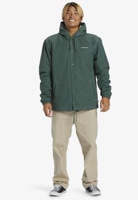Veste imperméable verte avec capuche, boutons-pression, poignets élastiques et ourlet à cordon de serrage, associée à un pantalon beige et des baskets noires.