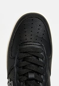 Gros plan sur le bout texturé noir d'une chaussure de sport avec des perforations et des lacets noirs sur une semelle blanche, photographié sur un fond clair.