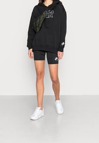 Person som bär en svart Nike Air-hoodie, svarta Nike Air-shorts, vita sneakers och en kamouflagemönstrad crossbody-väska framför en enhetlig bakgrund.