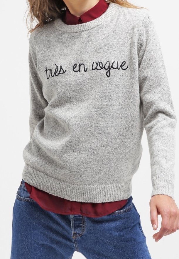 Personne portant un pull gris avec le texte « très en vogue », superposé sur une chemise bordeaux et associé à un jean bleu.