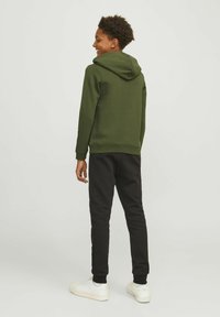 Felpa con cappuccio verde scuro con polsini a coste, abbinata a pantaloni della tuta neri e sneakers bianche. Il tessuto sembra morbido e informale.