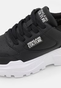 Baskets Versace Jeans Couture noires avec semelle blanche épaisse, lacets noirs et logo sur la languette et le panneau latéral.