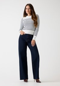Maglione a righe in bianco e blu navy, colletto alto con bottoni dorati, abbinato a pantaloni in denim a gamba larga blu scuro.