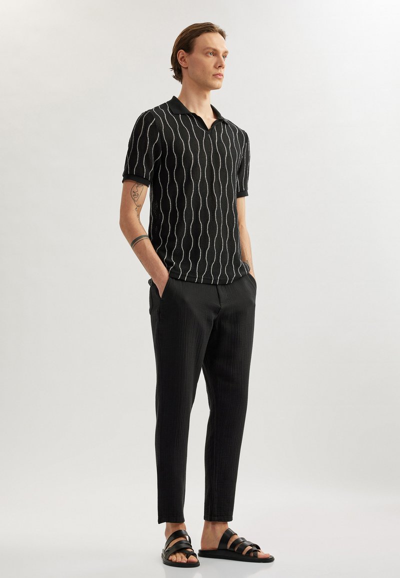 Schwarzes Strick-Polohemd mit weißen Wellenlinien, kombiniert mit schwarzen gestreiften Hosen und schwarzen Riemchensandalen. Glatte Textur, lässige Passform.
