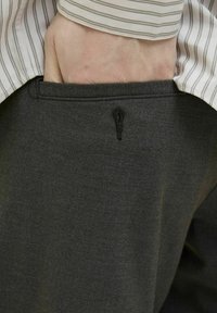 Pantaloni din țesătură gri închis, cu detalii de buzunar și o fante verticală. O cămașă în dungi bej și alb este parțial vizibilă deasupra.
