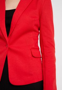 Blazer rouge en tissu texturé avec une fermeture à un bouton, des revers crantés et une poche latérale. Coupe ajustée avec des épaules structurées.
