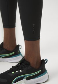 Leggings de sport noirs au design ajusté, présentant une texture lisse et une impression de logo. Associés à des chaussures de course noires et teal.