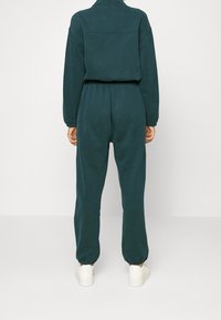 Dunkelblauer Jumpsuit aus weichem Stoff, mit hohem Kragen, elastischem Bund und schmal zulaufenden Beinen. Weiße Sneaker ergänzen den Look.