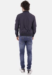 Mand vendt væk iført en mørk jakke, blå jeans og hvide sneakers mod en ensartet lys baggrund.