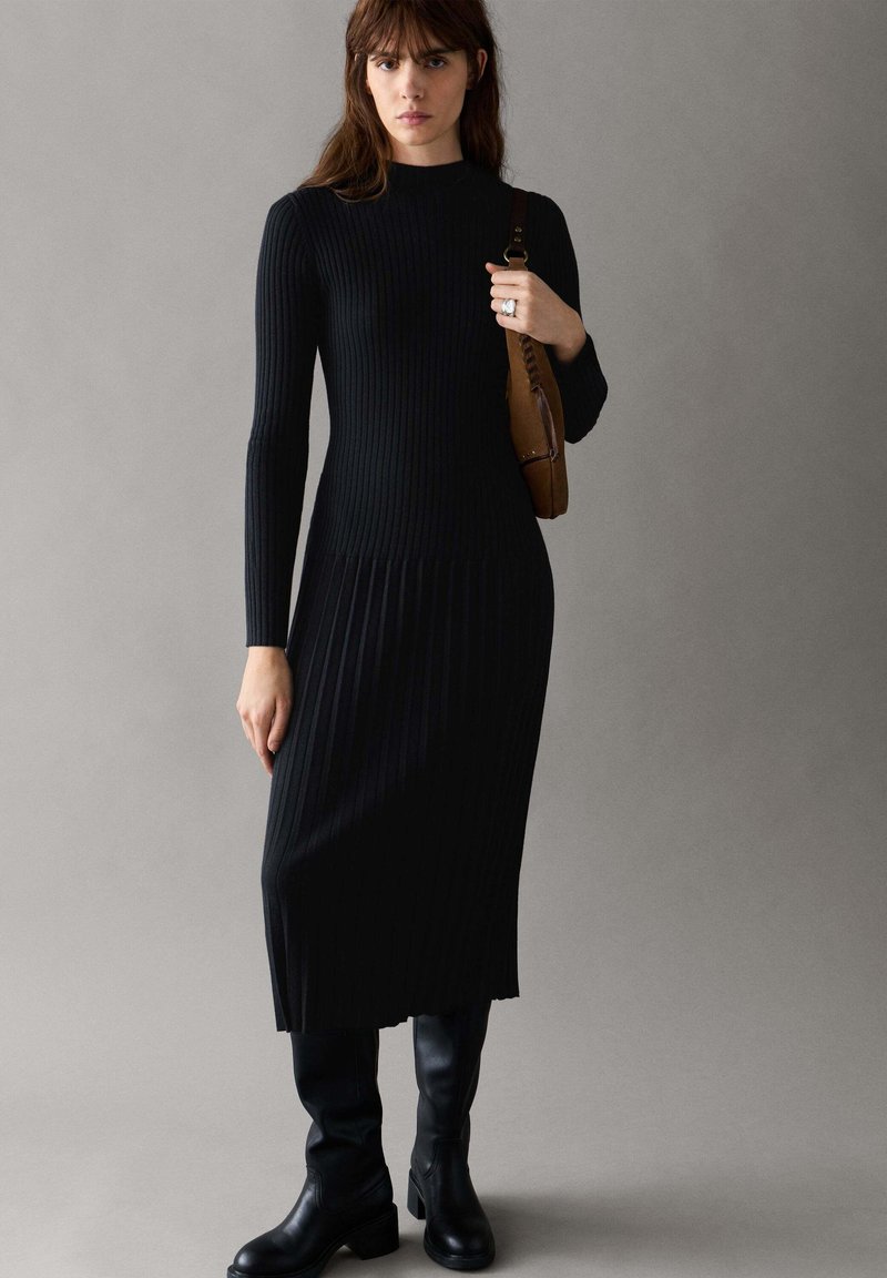 Femme debout portant une longue robe noire côtelée à manches longues et des bottes noires montant aux genoux, tenant un sac à bandoulière marron sur fond gris.