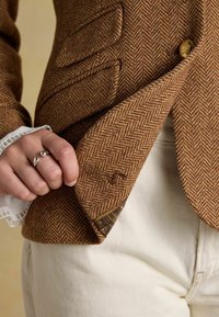 Blazer maro în model herringbone cu o suprafață texturată, având un buton decorativ și un căptușeală din material contrastant vizibilă în interiorul buzunarului.