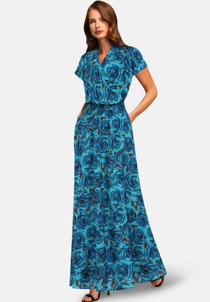 Žena stojaca s rukami v kapsách, oblečená v modrých kvetovaných maxi šatách a čiernych lodičkách s otvorenou špičkou na bielom pozadí.