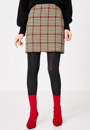 Femme portant un pull noir, une jupe à carreaux beige et rouge, des collants noirs et des bottines rouges à talons, debout sur un fond blanc.
