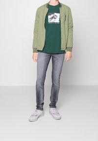 Chaqueta bomber acolchada verde, camiseta gráfica verde oscuro, jeans gris deslavado y zapatillas blancas con acentos rojos. Un conjunto simple y casual.