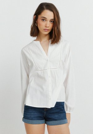 Młoda kobieta o brązowych włosach do ramion, ubrana w białą haftowaną bluzkę z długim rękawem oraz ciemnoniebieskie podwinięte dżinsowe szorty.