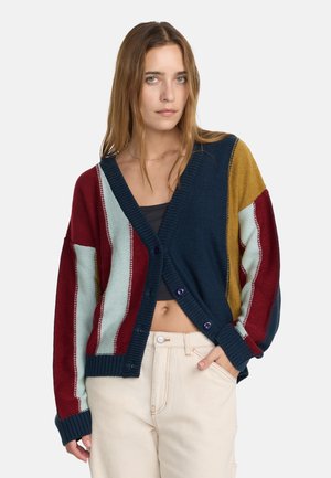Jeune femme portant un cardigan en maille multicolore avec des rayures bleu marine, rouge, bleu clair et jaune moutarde, associé à un pantalon beige clair.