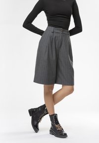 Shorts grigi a righe sottili con vita alta, abbinati a stivali neri con lacci e dettagli metallici. Visibile una maglietta nera a maniche lunghe.