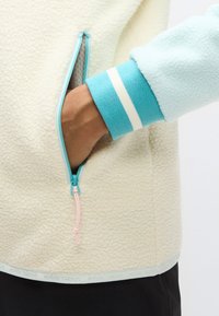 Ζακέτα από φλις σε κρεμ χρώμα με τυρκουάζ φερμουάρ και μανσέτες. Διαθέτει ανοιχτή τσέπη με ροζ ταινία έλξης και λευκές ρίγες στις μανσέτες.