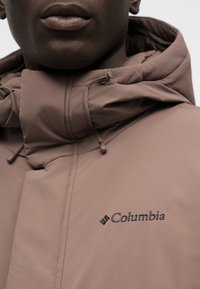Κοντινό πλάνο ατόμου που φορά καφέ μπουφάν Columbia με ρυθμιζόμενη κουκούλα και λογότυπο στο στήθος.