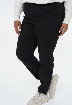 Pantalons noirs ajustés en denim avec une taille élastique, des poches latérales et une texture lisse, accompagnés de baskets claires.