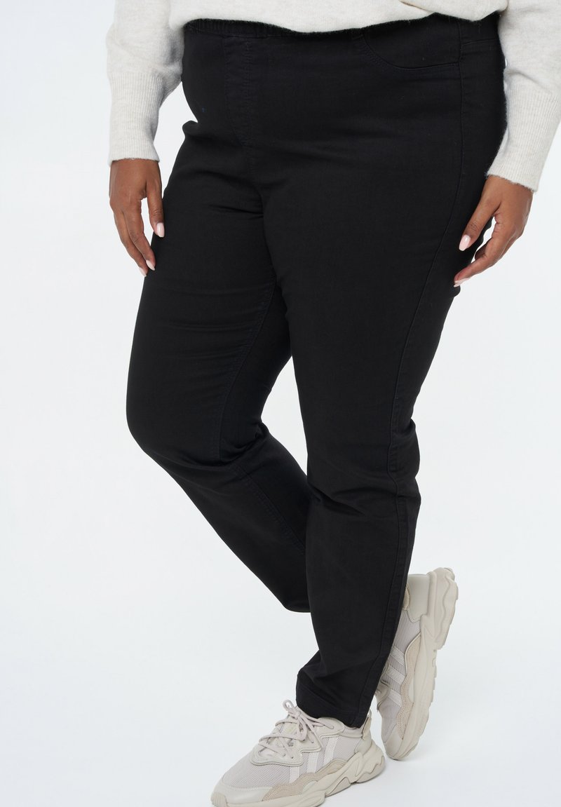 Pantalons noirs ajustés en denim avec une taille élastique, des poches latérales et une texture lisse, accompagnés de baskets claires.