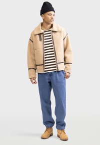 Beige nepbont jack met bruine accenten, gestreept shirt, blauwe spijkerbroek en tan werkschoenen. Model draagt een zwarte beanie.