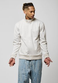 Chaqueta de cremallera beige claro con tejido texturizado, cuello alto y dos bolsillos laterales. Combinada con jeans claros desgastados.