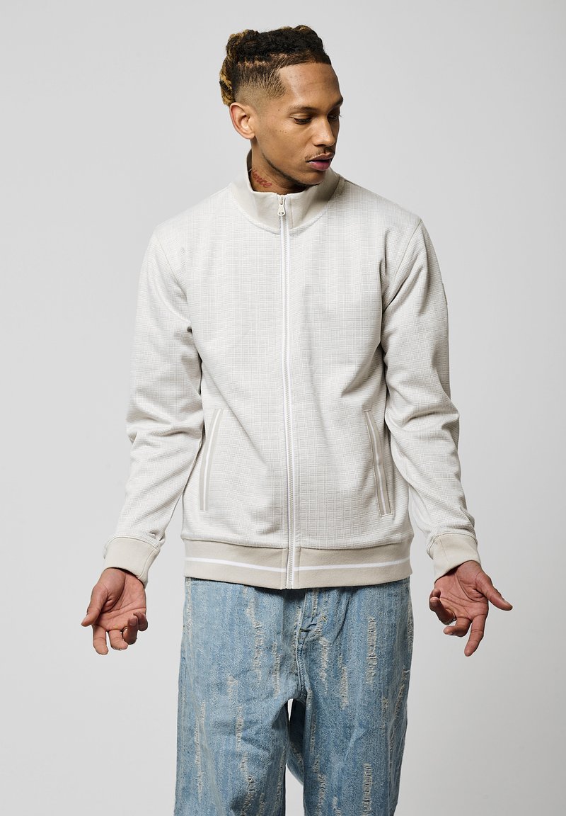 Chaqueta de cremallera beige claro con tejido texturizado, cuello alto y dos bolsillos laterales. Combinada con jeans claros desgastados.