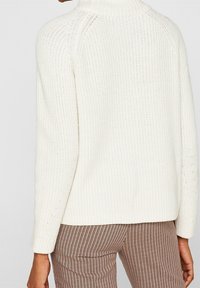 Pull blanc en maille texturée avec un col montant et des manches raglan. Des côtes visibles et des détails en maille ajourée mettent en valeur le design.
