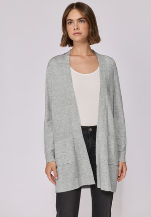 Cardigan grigio chiaro, con apertura frontale, texture morbida, maniche lunghe e due tasche frontali. Indossato sopra un top bianco e pantaloni neri.