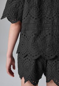 Sort stoftop med indviklet blomsterbroderi, udskårne mønstre og bølgede kanter. Præget mærkelogo på taljebåndet.