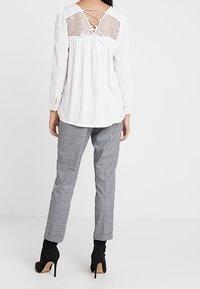 Top bianco a maniche lunghe con dettagli in pizzo sulla schiena, abbinato a pantaloni grigi a quadri e stivaletti neri con tacco alto.