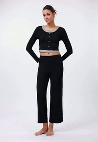 Νεαρή γυναίκα που στέκεται ξυπόλητη, φορώντας μαύρα παντελόνια με φαρδιά πόδια και ένα μαύρο κοντό πουλόβερ με κουμπιά πάνω από ένα γκρι μπλουζάκι, σε απλό φόντο.