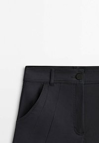 Gros plan sur la ceinture d’un pantalon noir avec passants de ceinture, fermeture à bouton et poche avant sur fond blanc uni.