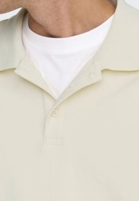 Polo de crema claro con cuello doblado, diseño liso, mangas cortas y un solo botón en el cuello, superpuesto a un cuello redondo blanco.
