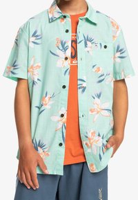 Blumiges kurzärmliges Hemd aus hellgrünem Stoff mit großen rosa und weißen Blumen, schwarzen Knöpfen und einer Brusttasche.