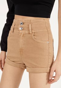 Pantalones cortos de mezclilla beige de tiro alto con dos botones frontales, puños enrollados y costuras de doble puntada. Presentan bolsillos delanteros y una textura suave.
