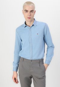 Chemise en denim bleu clair avec un col boutonné, des manches longues et un petit logo brodé. Portée avec un pantalon gris.