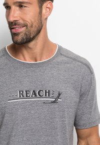 Graues T-Shirt mit kurzen Ärmeln aus weicher Baumwolle, mit schwarzem "REACH"-Druck und dekorativen Akzenten, rundem Halsausschnitt und dezenten Stichdetails.