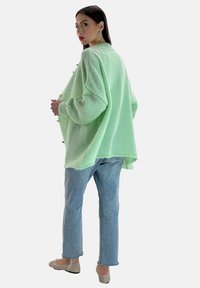 Miętowy, oversize'owy sweter z teksturowanym splotem, z obniżonymi ramionami i tyłem dłuższym od przodu, zestawiony z jasnymi dżinsami i neutralnymi balerinami.