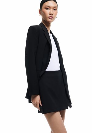 Vrouw draagt zwarte blazer en rok met wit topje, staande tegen een witte achtergrond, kijkt recht vooruit met een neutrale gezichtsuitdrukking.