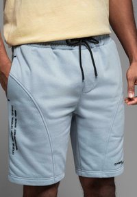 Lyseblå fleece shorts med elastisk talje, sort snor, sidesømme og minimalistisk tekstdetalje på venstre ben.