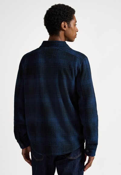 Muškarac okrenut leđima nosi tamnoplavu i crnu kariranu košulju dugih rukava i tamne traperice, stoji na jednobojnoj bijeloj pozadini.