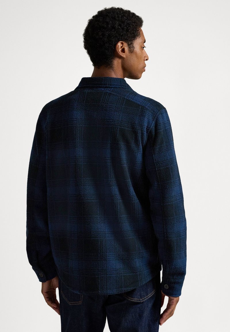 Mann von hinten, trägt ein dunkelblau-schwarz kariertes Langarmhemd und dunkle Jeans vor einem schlichten weißen Hintergrund.