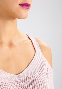 Pull en maille rose clair avec un col rond, présentant un motif texturé et des bretelles larges. Teint de peau lisse visible en arrière-plan.