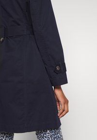 Manche de manteau bleu marine foncé avec poignet boutonné, portée sur la main, et pantalon imprimé léopard dans des tons bleus, gris et noirs.