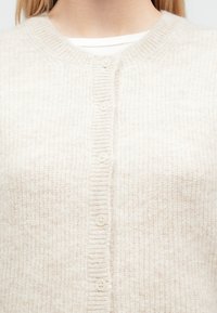 Bézs színű kötött kardigán kerek nyakkivágással, bordázott szegéllyel és hat világos színű gombbal az elején. Puha textúra és egyszerű kialakítás.