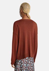 Kupferfarbener Strickpullover mit lockerer Passform, geripptem Saum und leicht überschnittenen Schultern, über einem blumenmusterbedruckten Kleid getragen.