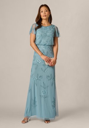 Vrouw draagt een lichtblauwe jurk tot op de grond met bloemenborduurwerk, doorschijnende korte mouwen, zilveren hakken en houdt een kleine beige clutch vast.